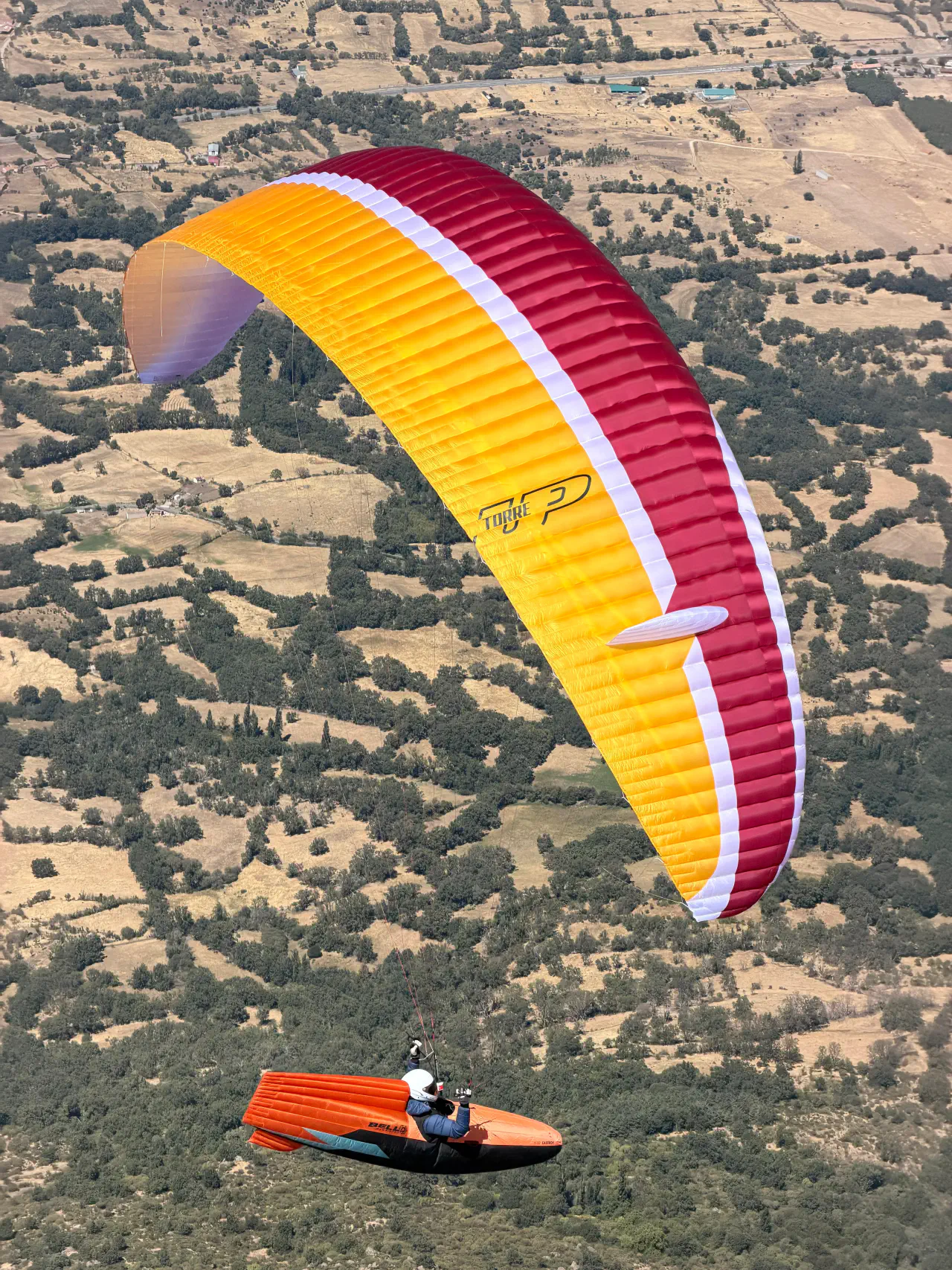A paraglider pilot in an orange pod harness flying a yellow, white and red wing above a patchwork of fields and trees